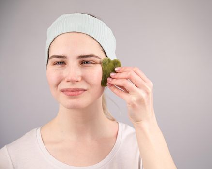 Portrait of a young woman massages her face with a gouache scraper on a white background