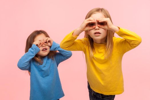 Adorable, curious, nosy two little girls looking through fingers imitating binoculars, exploring world, discovering something new with interested gaze. indoor studio shot isolated on pink background
