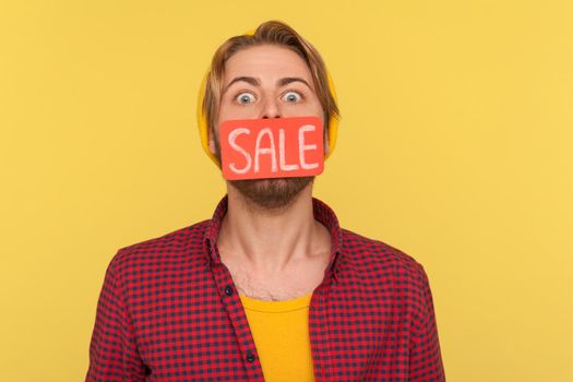 Amazed trendy hipster guy standing with Sale word on his mouth, looking at camera big eyes surprised, shocked by discounts, Black Friday low prices. indoor studio shot isolated on yellow background