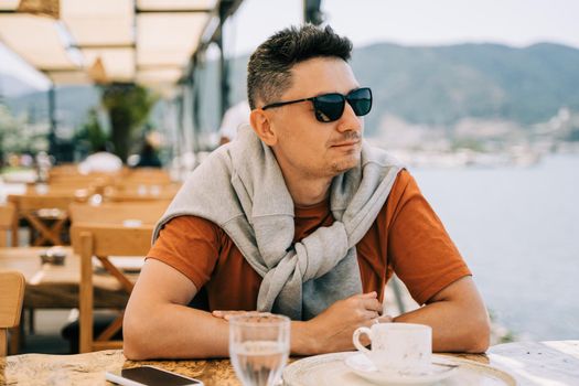 Young man male in sunglasses drinking coffee in a street outdoors seaside cafe restaurant coffee shop with scenery sea and mountains in the background. Hello summer holiday vacation.