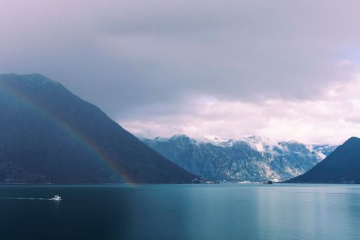 A double rainbow stretches across the sea water of Kotor Bay, Montenegro, surrounded by dramatic clouds and snow-capped mountains, with a boat speeding into view