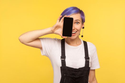 Portrait of happy excited hipster girl with violet short hair in denim overalls covering half face with cell phone and looking at camera with toothy smile. isolated on yellow background, studio shot