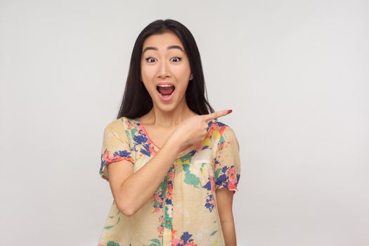 Look, awesome advertise here, Fascinating asian girl with brunette hair in summer blouse pointing aside and looking with shocked expression, showing copy space, empty place for commercial. studio shot