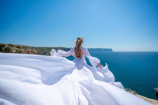 Blonde with long hair on a sunny seashore in a white flowing dress, rear view, silk fabric waving in the wind. Against the backdrop of the blue sky and mountains on the seashore