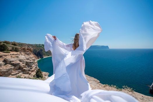 Blonde with long hair on a sunny seashore in a white flowing dress, rear view, silk fabric waving in the wind. Against the backdrop of the blue sky and mountains on the seashore