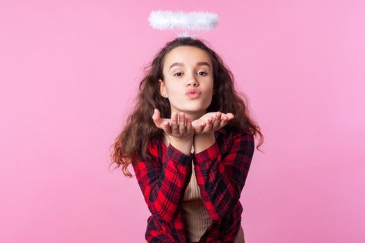 Portrait of lovely teenage girl with curly brunette hair in plaid shirt and toy halo above head sending air kiss with pouted lips, romance on valentines day. studio shot isolated on pink background