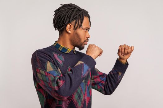 Profile portrait serious aggressive african man with dreadlocks holding clenched fists up ready to boxing, defending his rights and freedom. Indoor studio shot isolated on gray background