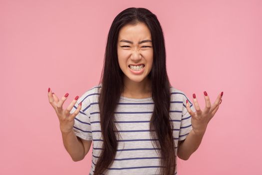 Portrait of enraged furious girl with long hair in striped t-shirt standing with clenched teeth and raised hands expressing wild anger, feeling crazy. indoor studio shot isolated on pink background