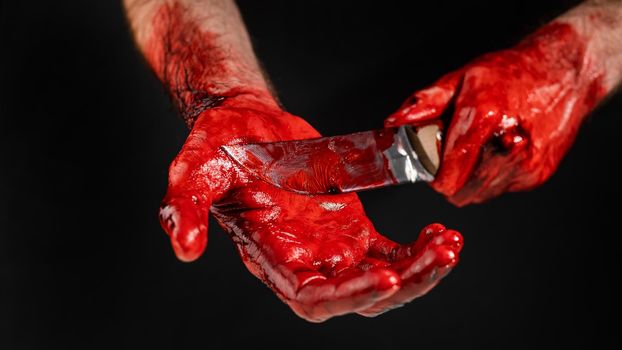 A man with blood-stained hands cuts his palm with a knife on a black background