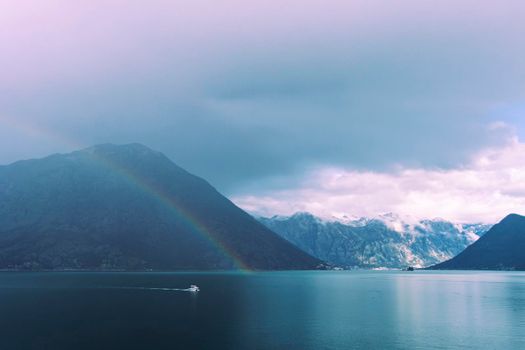 A double rainbow stretches across the sea water of Kotor Bay, Montenegro, surrounded by dramatic clouds and snow-capped mountains, with a boat speeding into view