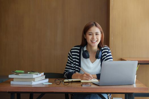 Young asian girl student wear headphone and writing a notebook and watching online class by laptop. e-learning education concept