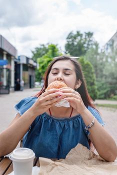 Summer vacation, street food eating. Charming hungry stylish woman, enjoying eating a burger outdoors, dressed in jeans shirt, wearing sunglasses