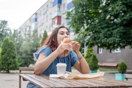 Summer vacation, street food eating. Charming hungry stylish woman, enjoying eating a burger outdoors, dressed in jeans shirt, wearing sunglasses