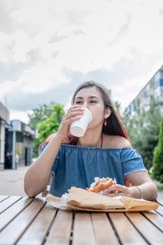 Summer vacation, street food eating. Charming hungry stylish woman, enjoying eating a burger outdoors, dressed in jeans shirt, wearing sunglasses