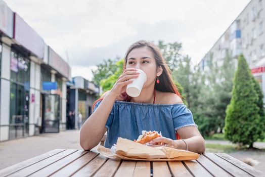 Summer vacation, street food eating. Charming hungry stylish woman, enjoying eating a burger outdoors, dressed in jeans shirt, wearing sunglasses