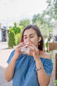 Summer vacation, street food eating. Charming hungry stylish woman, enjoying eating a burger outdoors, dressed in jeans shirt, wearing sunglasses