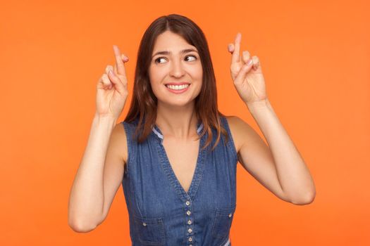 Cute positive brunette woman smiling happily and holding fingers crossed for good luck, hoping for fortune, success, making wish of something girlish pleasant. indoor studio shot orange background