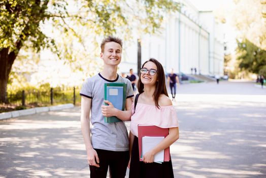 Portrait of two happy students standing in park near university with books, exercise books, folders before classes in good mood smiling to camera