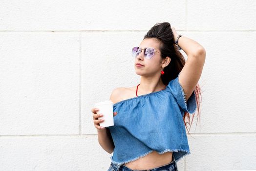 coffee to go. portrait of stylish young woman wearing jeans shirt, sunglasses and bag, drinking coffee to go at street, white wall background