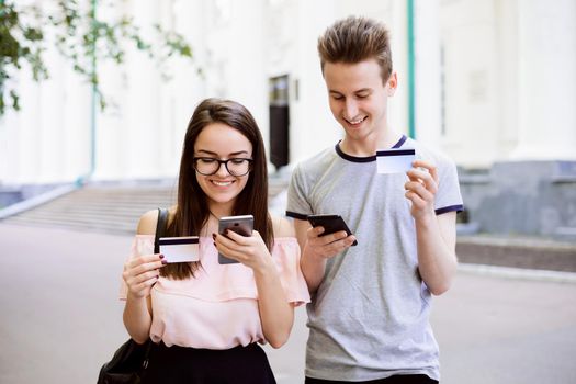 Attractive male and female standing in front of conventional university with credit cards and mobile phones in hands, purchasing things in the web