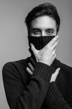 Portrait of stylish handsome young man in warm sweater, covering mouth with high collar and looking pensive thoughtful, male fashion, vogue concept. black and white, indoor studio shot isolated