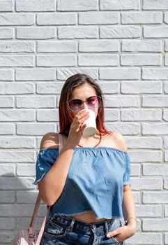 Summer vacation, street food eating. Charming stylish woman, enjoying drinking coffee outdoors, dressed in jeans shirt, wearing sunglasses on white brick wall background