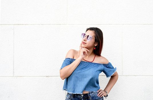 stylish young woman wearing jeans shirt, sunglasses and bag, at street, white wall background, summer look copy space