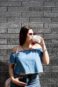 coffee to go. stylish young woman wearing jeans shirt, sunglasses and bag, drinking coffee to go at street, black brick wall background