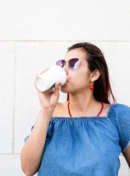 coffee to go. portrait of stylish young woman wearing jeans shirt, sunglasses and bag, drinking coffee to go at street, white wall background