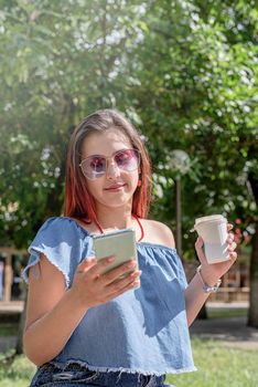 Attractive young woman in summer clothes and sunglasses holding cup of coffee in her hands and using smartphone videocall while sitting on bench in the park