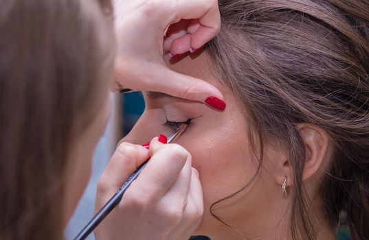 Close-up of the makeup artist's hands draw arrows on the eyes with a brush. Female master makes makeup to a young woman. Business concept - beauty salon, facial skin care, cosmetology.