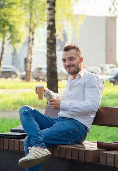 A serious man sits on a bench, drinks coffee, holds dollars. Young man on the background of passing cars, hot sunny summer day. Warm soft light, close-up.
