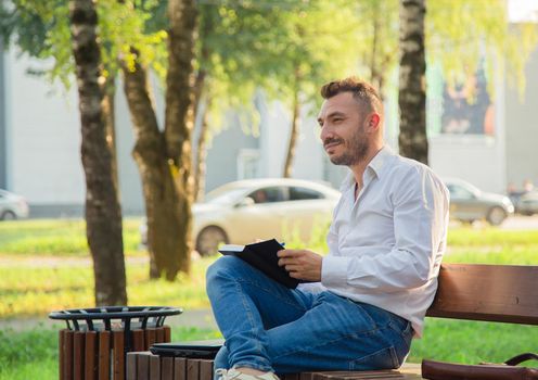 A happy man sits on a bench, makes plans, writes in a notebook. A young man on a background of green trees, a hot sunny summer day. Warm soft light, close-up.