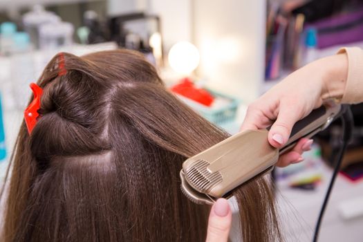 Close-up of a woman's hand straightens a lock of hair with a curling iron. The hairdresser makes a hairstyle for a young woman. Barber shop, business concept. Beauty salon, hair care.