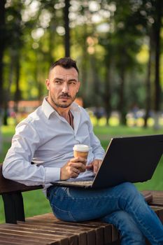 Serious man works in the park with a laptop, drinks coffee. A young man on a background of green trees, a hot sunny summer day. Warm soft light, close-up.