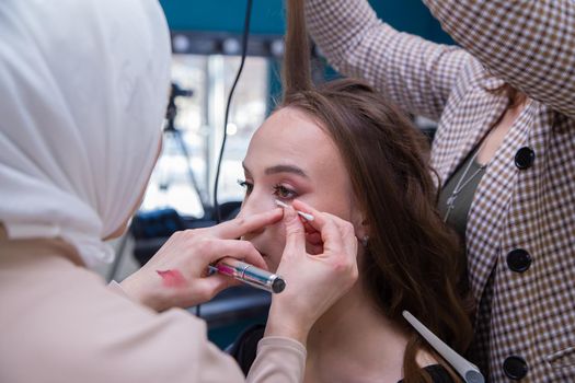 Women's hands paint the girl's eyelids and curl her hair in the salon. Make-up artist and hairdresser create an image of a young woman. Business concept - beauty salon, facial skin and hair care.