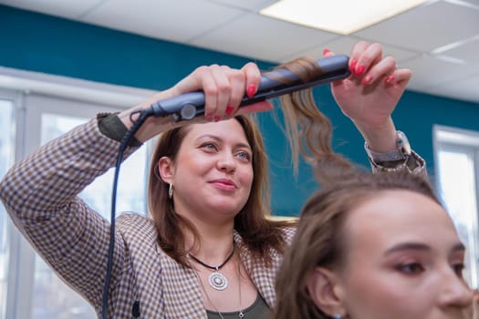 The girl master with a smile twists a lock of the client's hair on tongs. The hairdresser makes a hairstyle for a young woman. Barber shop, business concept. Beauty salon, hair care.