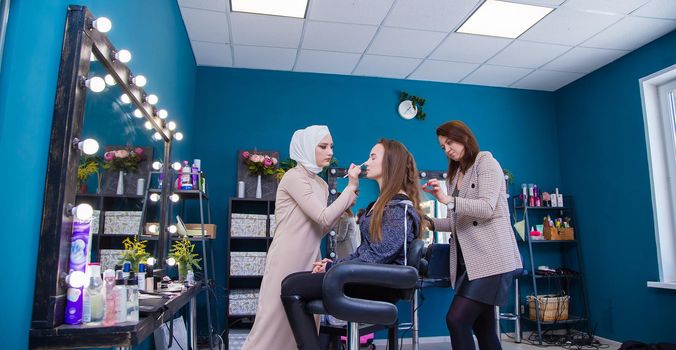 Two masters style the girl's hair and paint her eyes with a brush. Make-up artist and hairdresser create an image of a young woman. Business concept - beauty salon, facial skin and hair care.