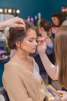 Women's hands paint the girl's eyes and curl her hair in the salon. Make-up artist and hairdresser create an image of a young woman. Business concept - beauty salon, facial skin and hair care.