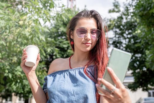 Attractive young woman in summer clothes and sunglasses holding cup of coffee in her hands,using smartphone videocall while sitting on bench in the park