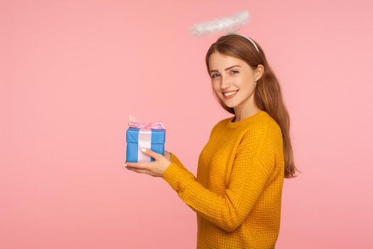 Charity and human kindness. Side view of angelic ginger girl with halo over head holding gift box and smiling at camera, giving holiday present to copy space. studio shot isolated on pink background