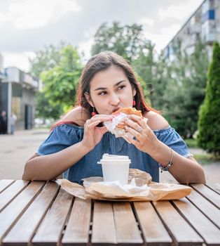 Summer vacation, street food eating. Charming hungry stylish woman, enjoying eating a burger outdoors, dressed in jeans shirt, wearing sunglasses