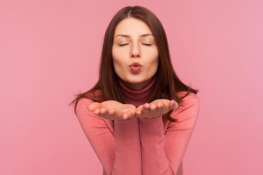 Beautiful tender brunette woman sending air kisses to camera, love and affection, saint valentines day celebration. Indoor studio shot isolated on pink background