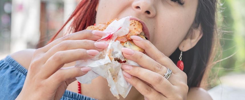 Summer vacation, street food eating. Charming hungry stylish woman, enjoying eating a burger outdoors, dressed in jeans shirt, wearing sunglasses