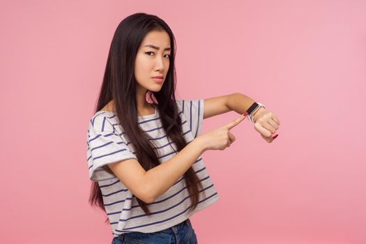 Hurry up, it's late. Portrait of displeased angry girl with long brunette hair pointing at wrist watch, checking time on clock and worrying about delay. indoor studio shot isolated on pink background