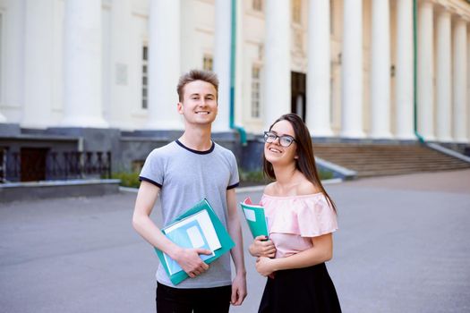 Portrait of two happy students standing in front of university before classes holding books, folders