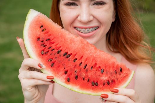 Beautiful red-haired woman smiling with braces and about to eat a slice of watermelon outdoors in summer.