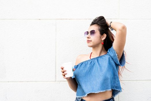 coffee to go. portrait of stylish young woman wearing jeans shirt, sunglasses and bag, drinking coffee to go at street, white wall background