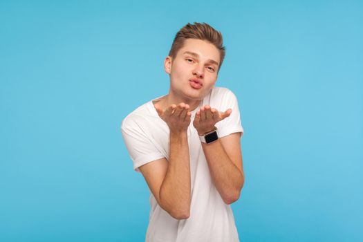 Portrait of handsome young man in white t-shirt sending sensual air kiss over palms to camera, demonstrating romantic feelings, love and affection. indoor studio shot isolated on blue background