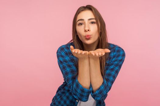 Portrait of charming girl in shirt sending sensual air kiss over palms, expressing tender romantic emotions and looking with love, flirting to camera. indoor studio shot isolated on pink background